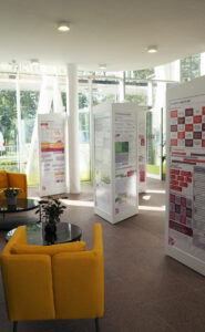 Infosäulen der Wanderausstellung Pfad der Vielfalt mit bunten Grafiken in einer Lounge mit Fenstern und Möbeln