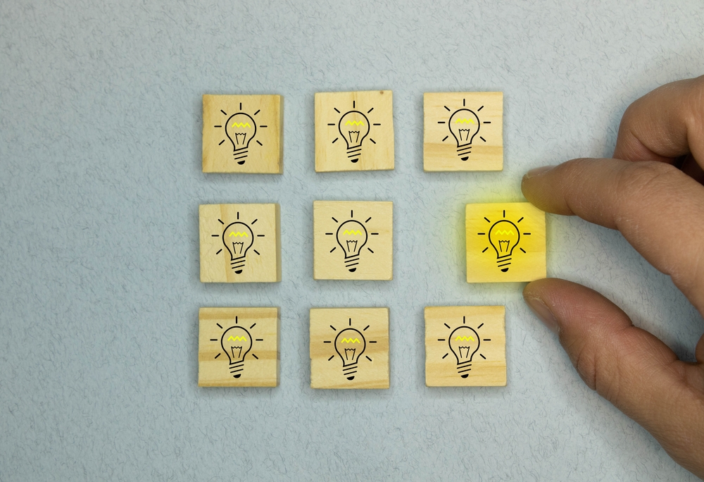 Eine Hand hebt ein Holzplättchen mit einer gelb leuchtenden Glühbirne an. Es befindet sich inmitten von acht weiteren Plättchen mit identischen, aber nicht leuchtenden Glühbirnenmotiven.